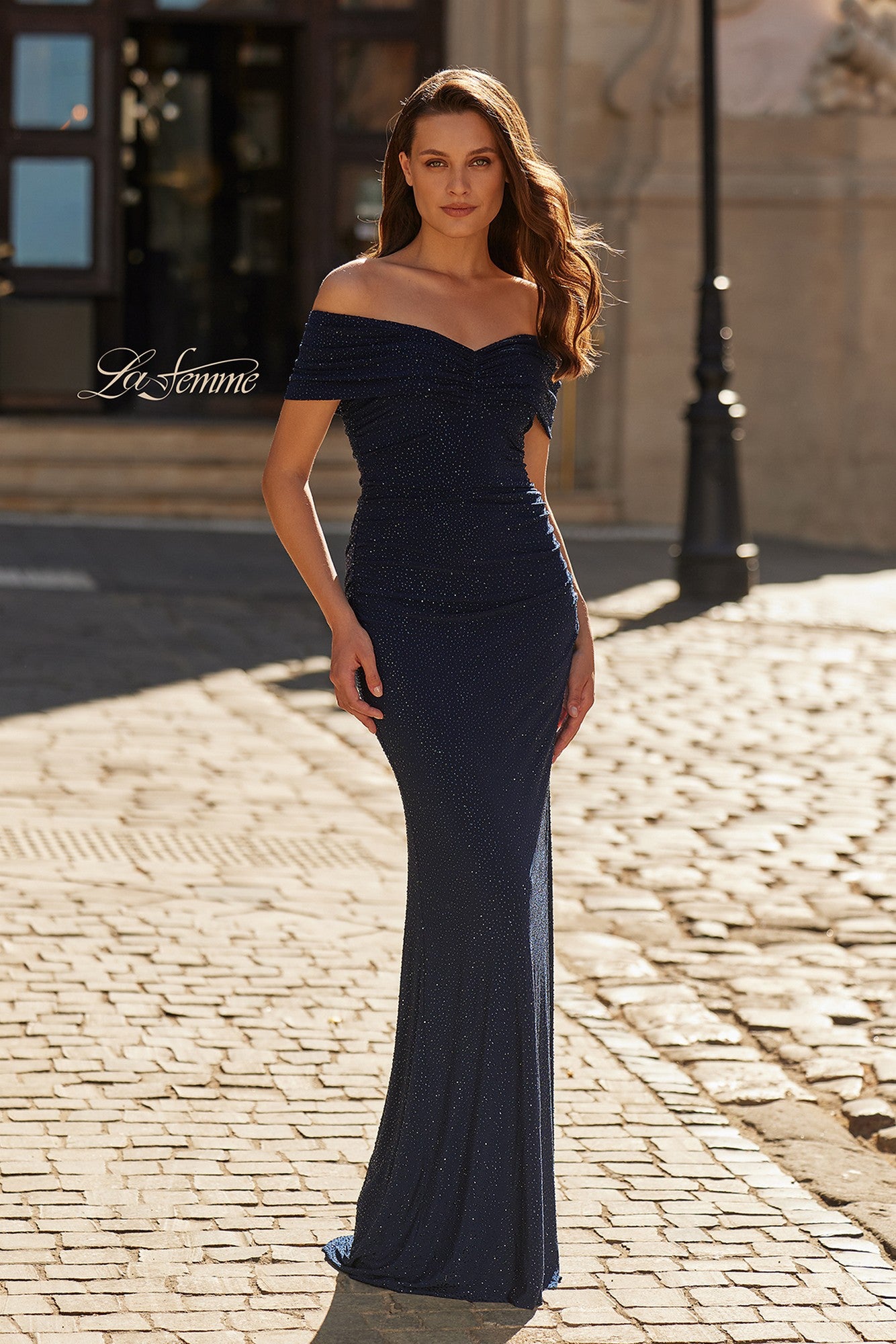 A woman in the La Femme 33312 long formal dress in navy stands on a sunlit cobblestone street, her floor-length gown elegantly complementing the scene.